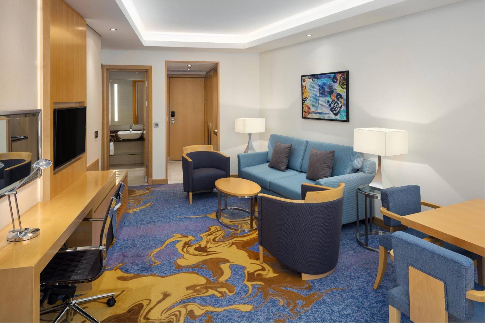 فندق شيراتون مكة جبل الكعبة المظهر الخارجي الصورة The photo displays a modern hotel room or suite. The room features a comfortable seating area with a blue couch and two blue armchairs surrounding a small round table. A wooden desk with a black office chair is positioned against one wall, accompanie