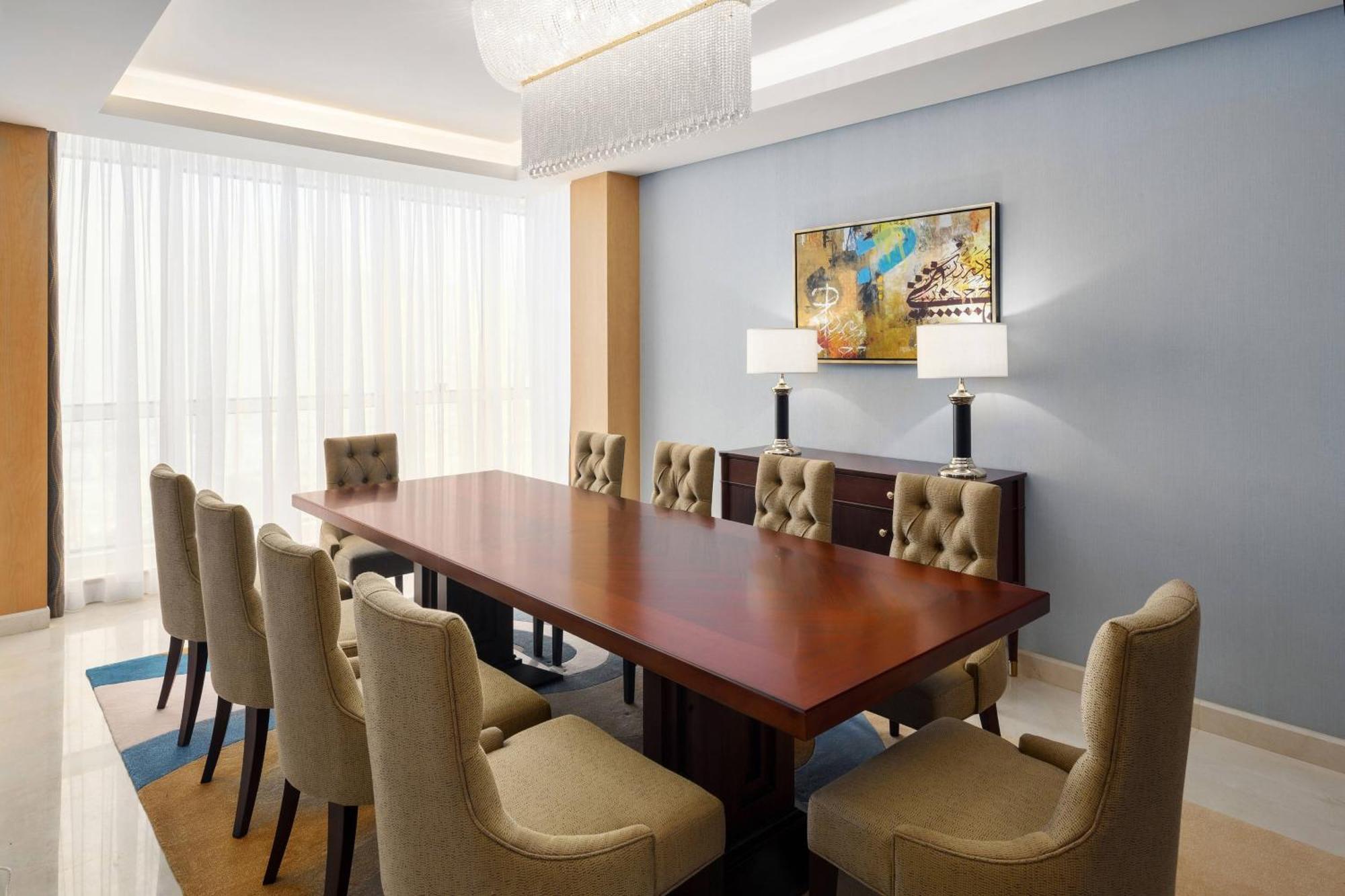 فندق شيراتون مكة جبل الكعبة المظهر الخارجي الصورة The photo shows a spacious and elegantly designed conference room. It features a large wooden conference table surrounded by upholstered chairs. The chairs have a beige color and a tufted design. On one side of the room, there are two contemporary la