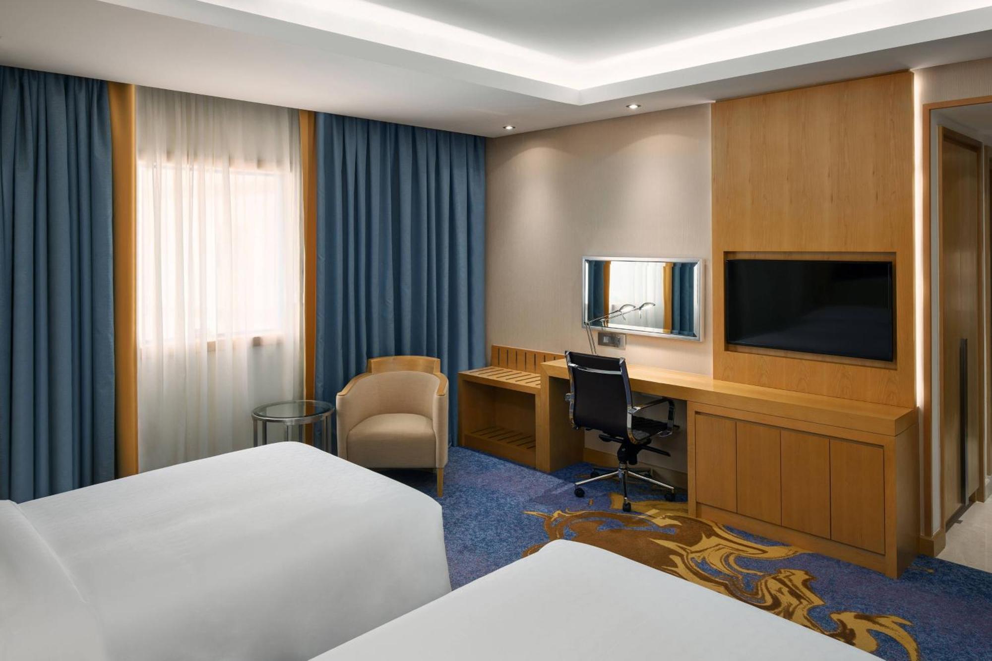 فندق شيراتون مكة جبل الكعبة المظهر الخارجي الصورة The photo shows a hotel room featuring two beds with white linens. There are large windows covered with blue curtains that allow natural light to enter. On one side of the room, there is a wooden workstation with a chair and a mirror, along with a fl