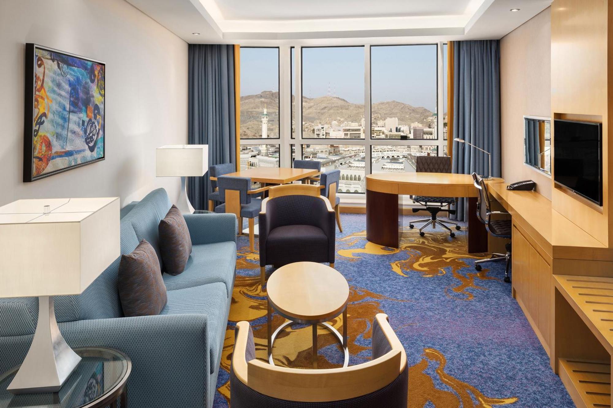 فندق شيراتون مكة جبل الكعبة المظهر الخارجي الصورة The photo shows a well-decorated hotel room featuring a modern and comfortable interior. There is a cozy seating area with a blue sofa and armchairs, complemented by a round coffee table. A desk with a chair is also present, ideal for work. The room 