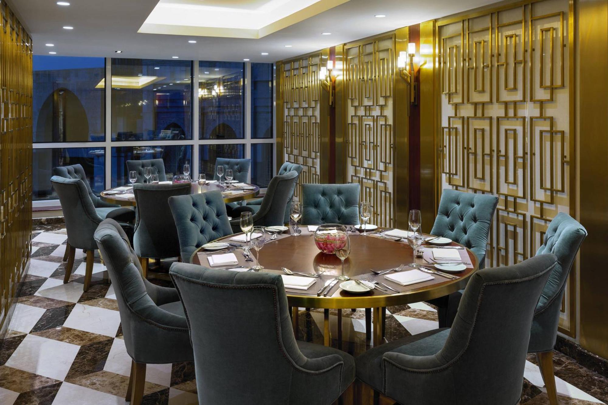 فندق شيراتون مكة جبل الكعبة المظهر الخارجي الصورة The photo shows an elegant dining room with a sophisticated ambiance. It features a round wooden table at the center, surrounded by plush chairs upholstered in a soft, teal fabric. The room has a stylish décor, highlighted by a geometrically patterne