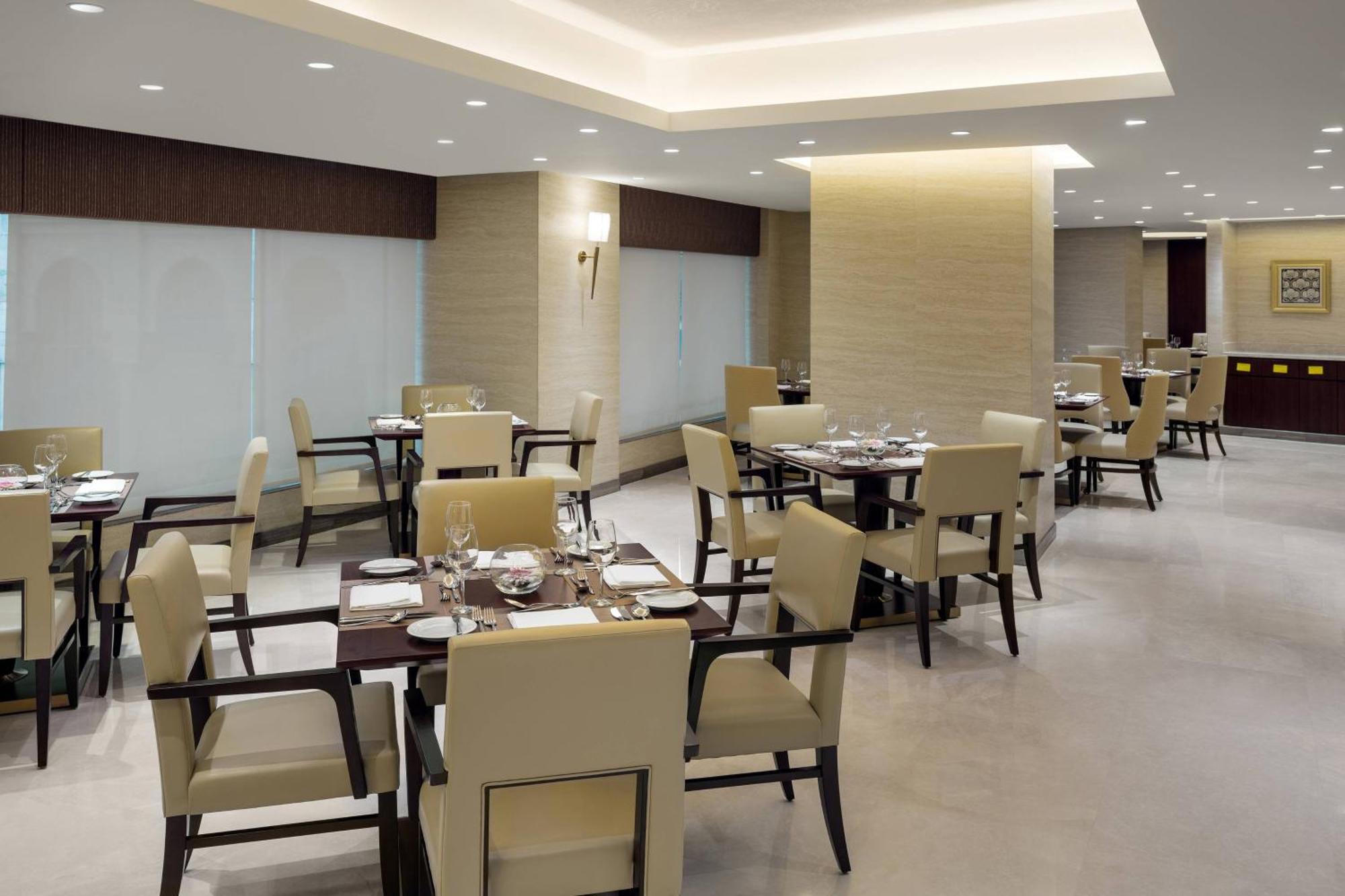 فندق شيراتون مكة جبل الكعبة المظهر الخارجي الصورة The photo shows a modern restaurant interior. It features several dining tables, each set with utensils and tableware. The chairs are upholstered in a light fabric, and the overall decor is sleek and minimalistic, with soft lighting from overhead fix