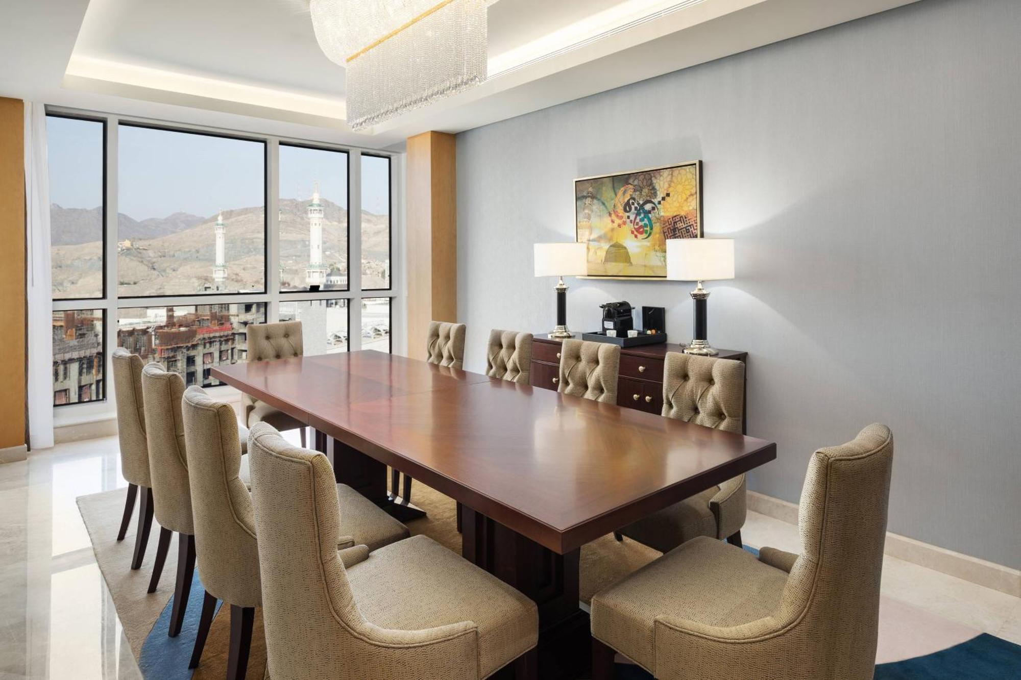 فندق شيراتون مكة جبل الكعبة المظهر الخارجي الصورة The photo shows a modern conference room with a large wooden table surrounded by upholstered chairs. The room is well-lit, featuring large windows that offer a view of the outdoors, possibly with mountains in the background. On one wall, there is a p